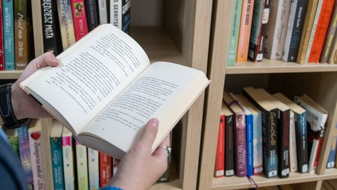 Biblioteczka przy ul. Objazdowej 17. Dłonie trzymające otwartą książkę na tle regału bibliotecznego.